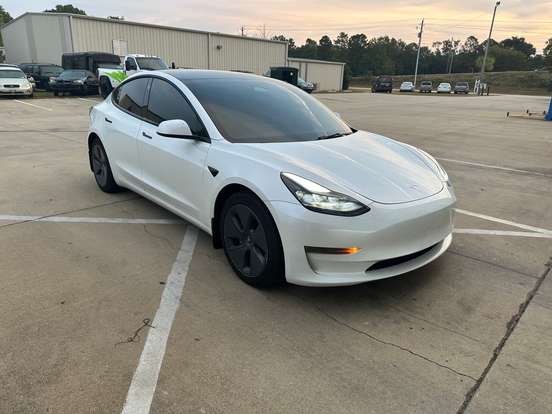 A white electric car parked in an outdoor parking lot near other vehicles and a building.