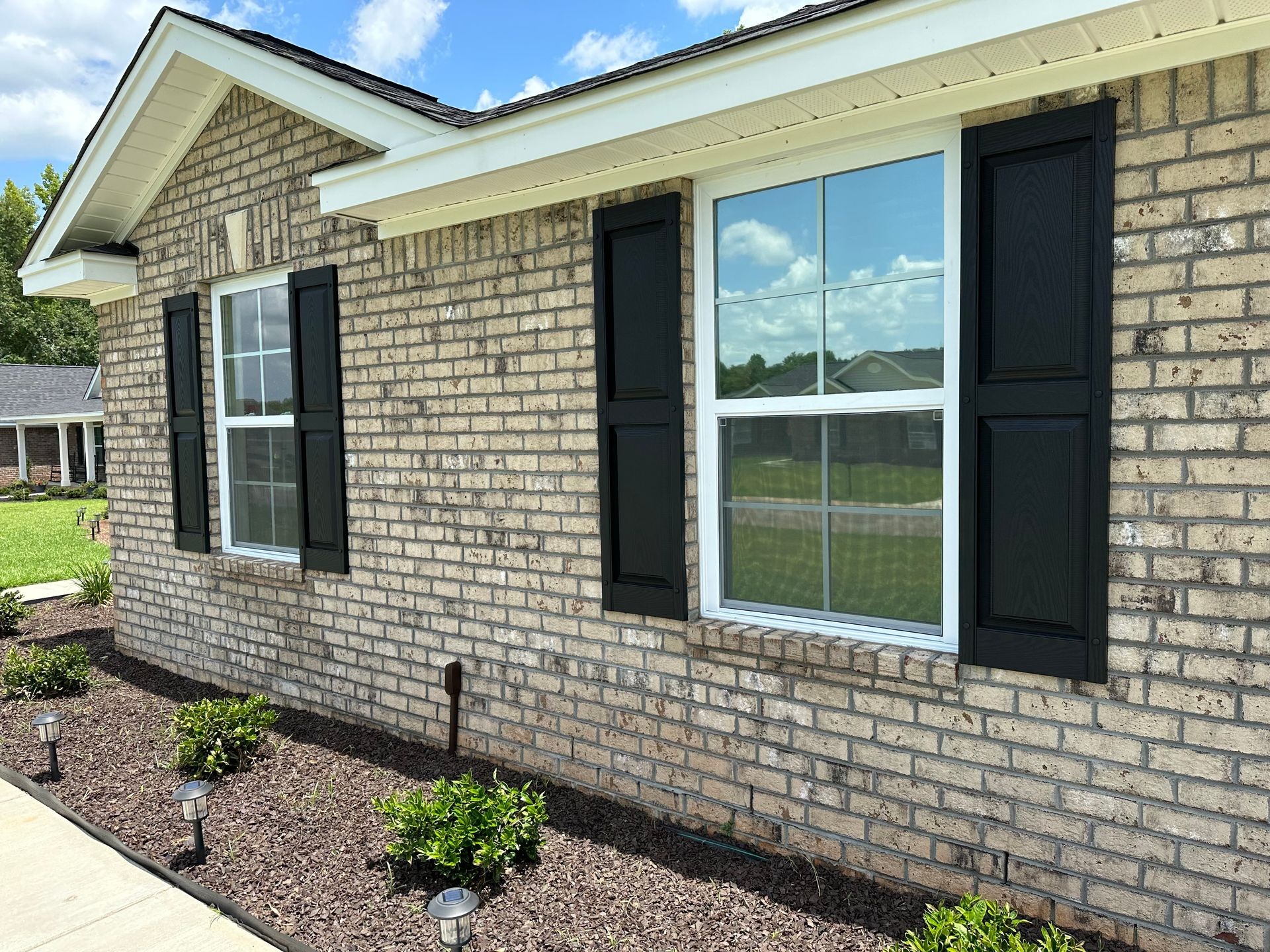 Exterior view of a brick house wall with black window shutters and landscaped flower bed.