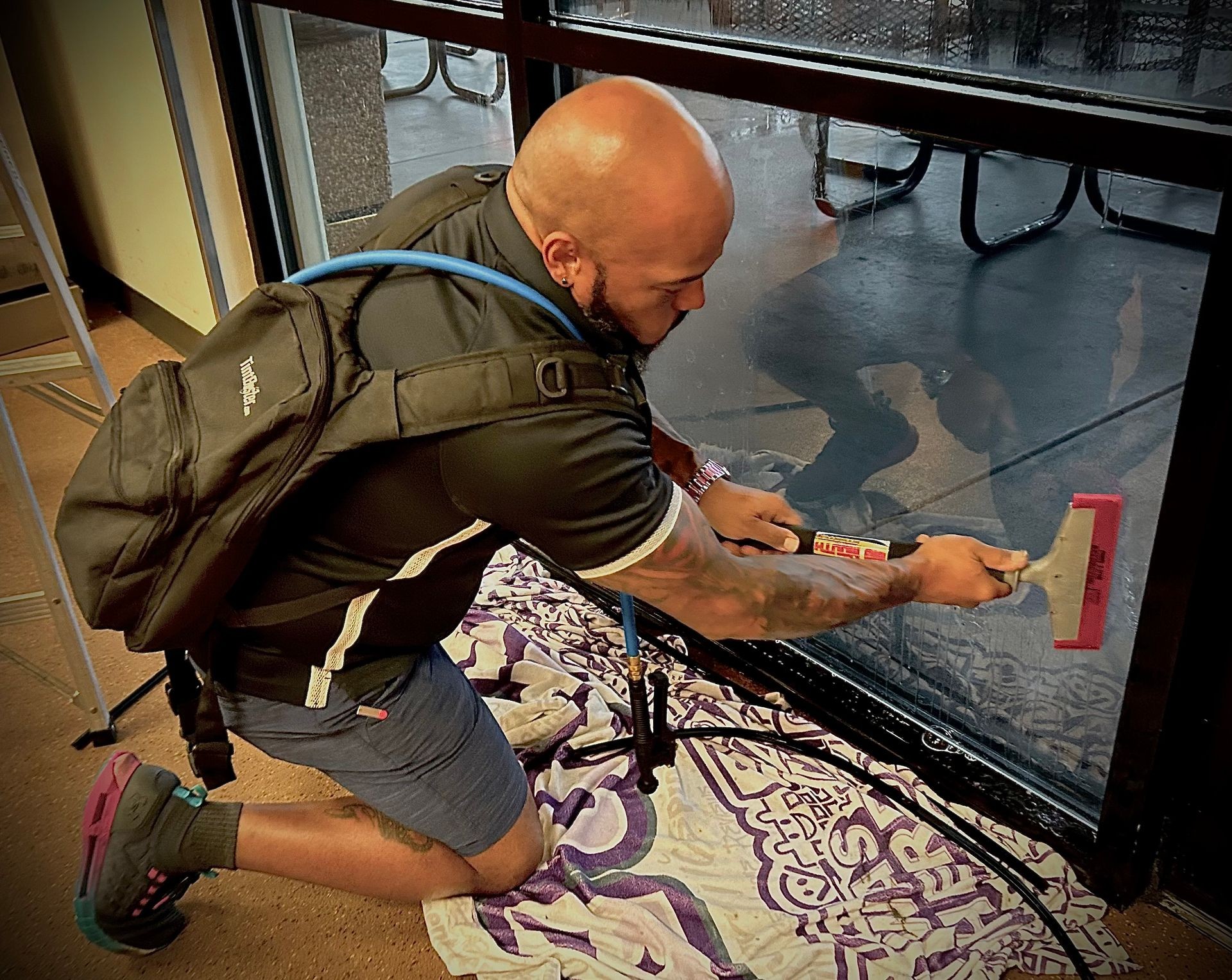 Person kneeling and cleaning a glass door with a squeegee, wearing a backpack indoors.